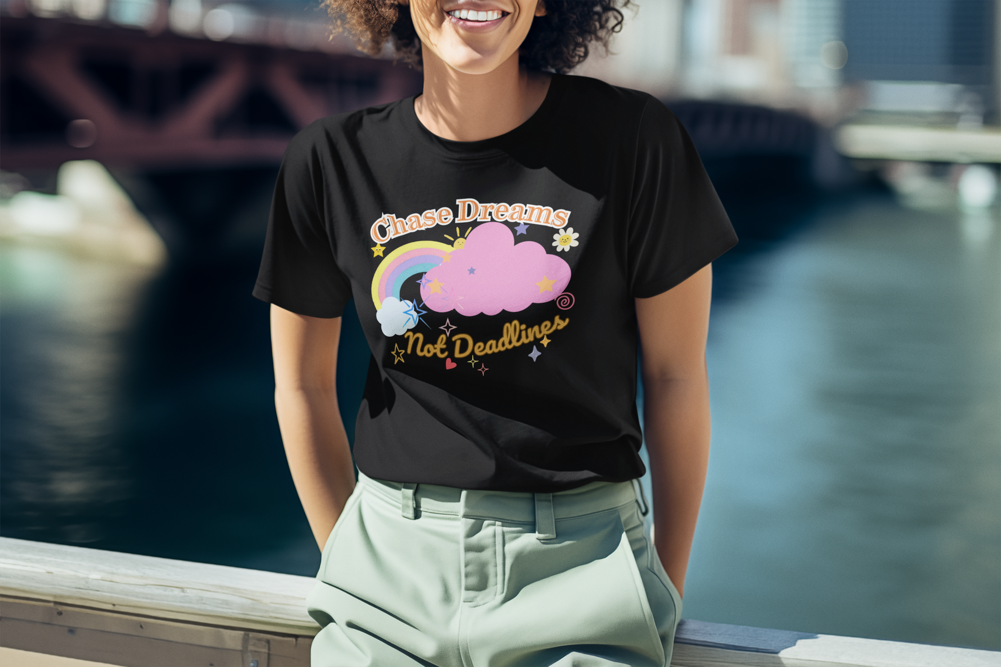 A woman wearing a black unisex graphic t-shirt with the phrase 'Chase Dreams, Not Deadlines' in bold lettering, featuring a rainbow and soft pink cloud